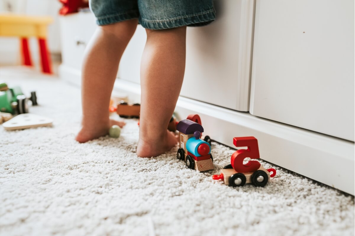 Toddler standing next to a shelf with wooden toys on the carpet next to him.