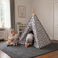 Two children playing with a gray star-patterned teepee tent indoors, one sitting with stuffed animals and the other crawling inside in a cozy playroom.