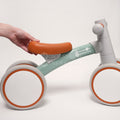 Hand holding the seat of a modern Montessori Generation baby balance bike, featuring a minimalist design with orange, mint green, and gray colors.