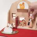 A close-up of a hand placing a wooden female farmer figure inside a Montessori-style wooden barn, surrounded by a swan in a pond, a dog, and a horse figure.